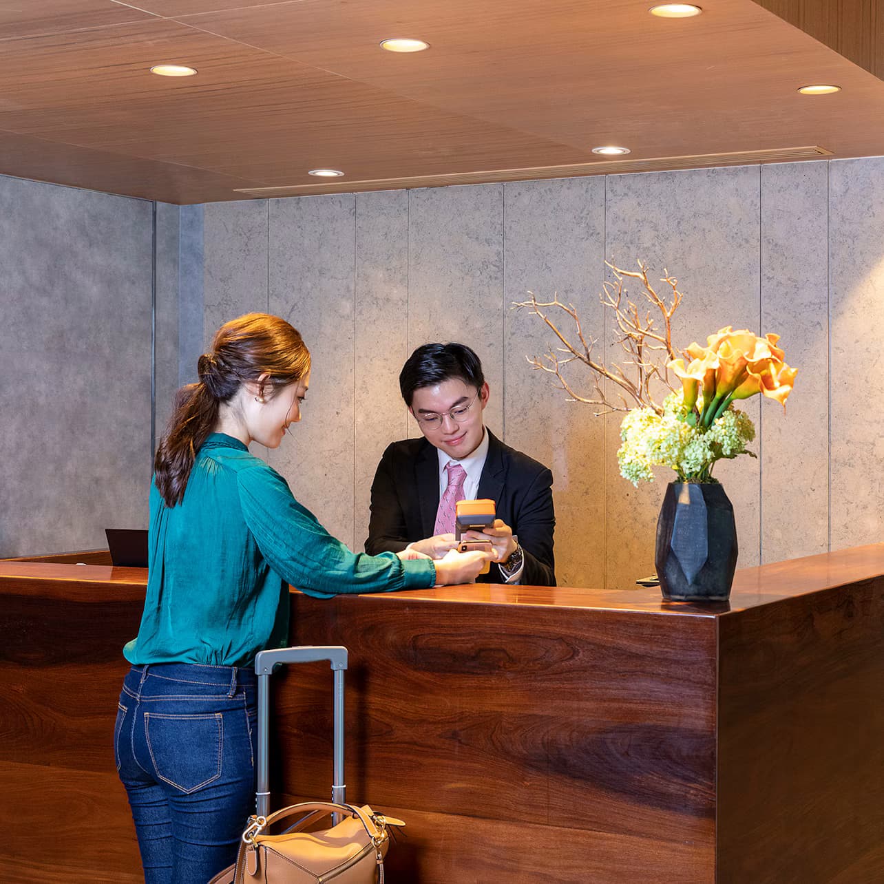 A woman is checking into a hotel at the reception desk, interacting with a male receptionist holding a card reader.