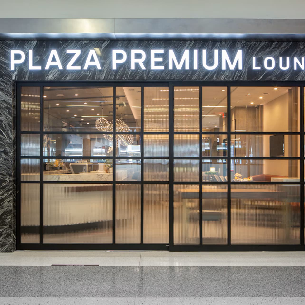 Entrance to Plaza Premium Lounge with illuminated sign, marble-patterned wall, large glass doors, and an information screen on the left.