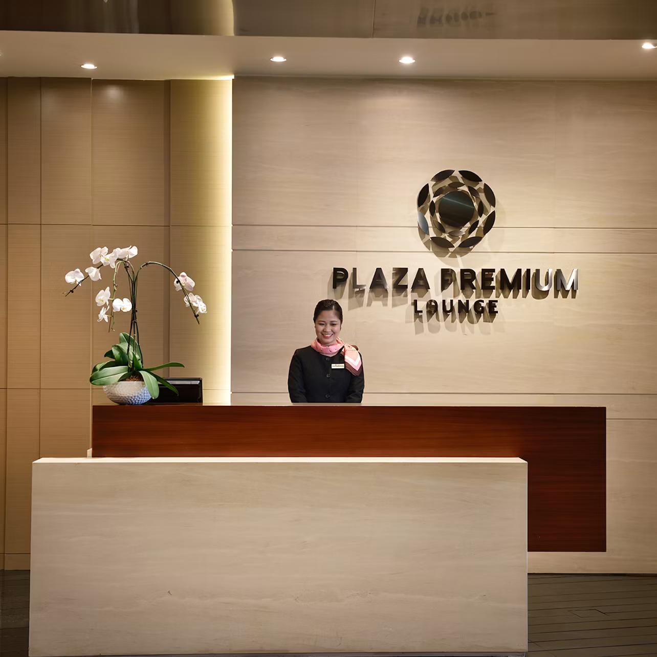 A reception desk with a friendly staff member, orchid plant, and "Plaza Premium Lounge" sign on the wall behind.