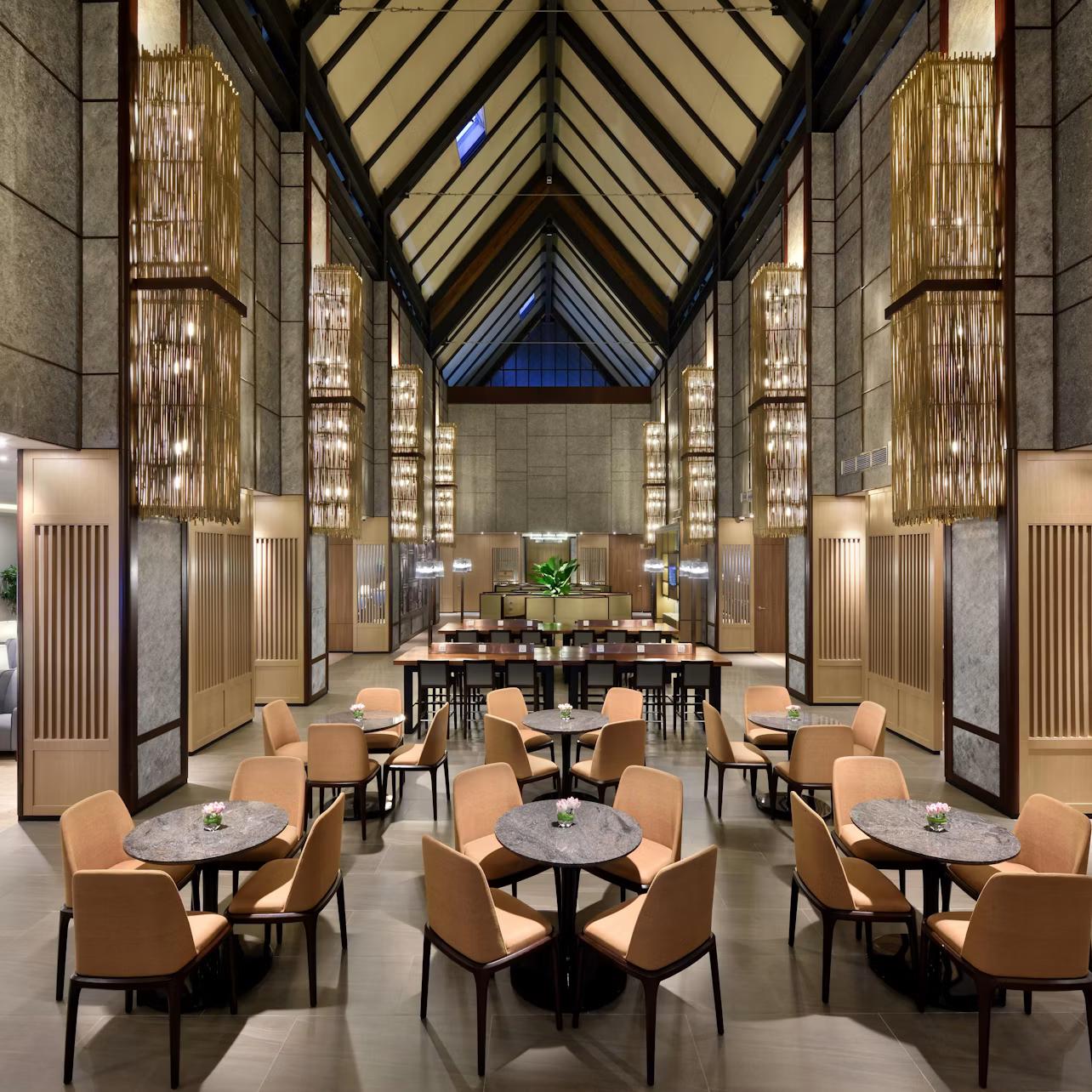 Spacious hotel lobby with vaulted ceiling, modern chandeliers, circular tables with beige chairs, and a central communal table.
