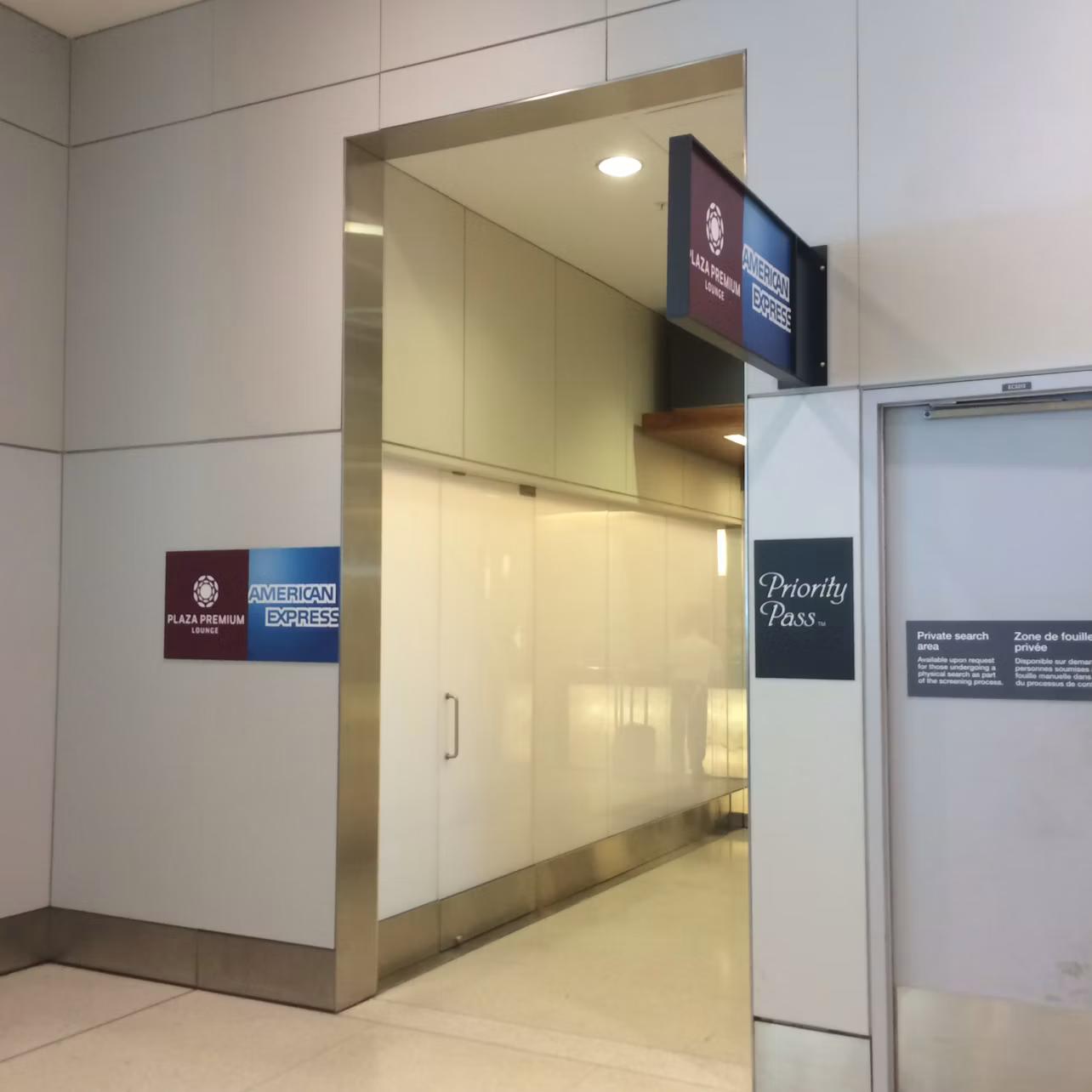 Entrance to an airport lounge with signs for Plaza Premium Lounge, American Express, and Priority Pass against a white and grey wall.
