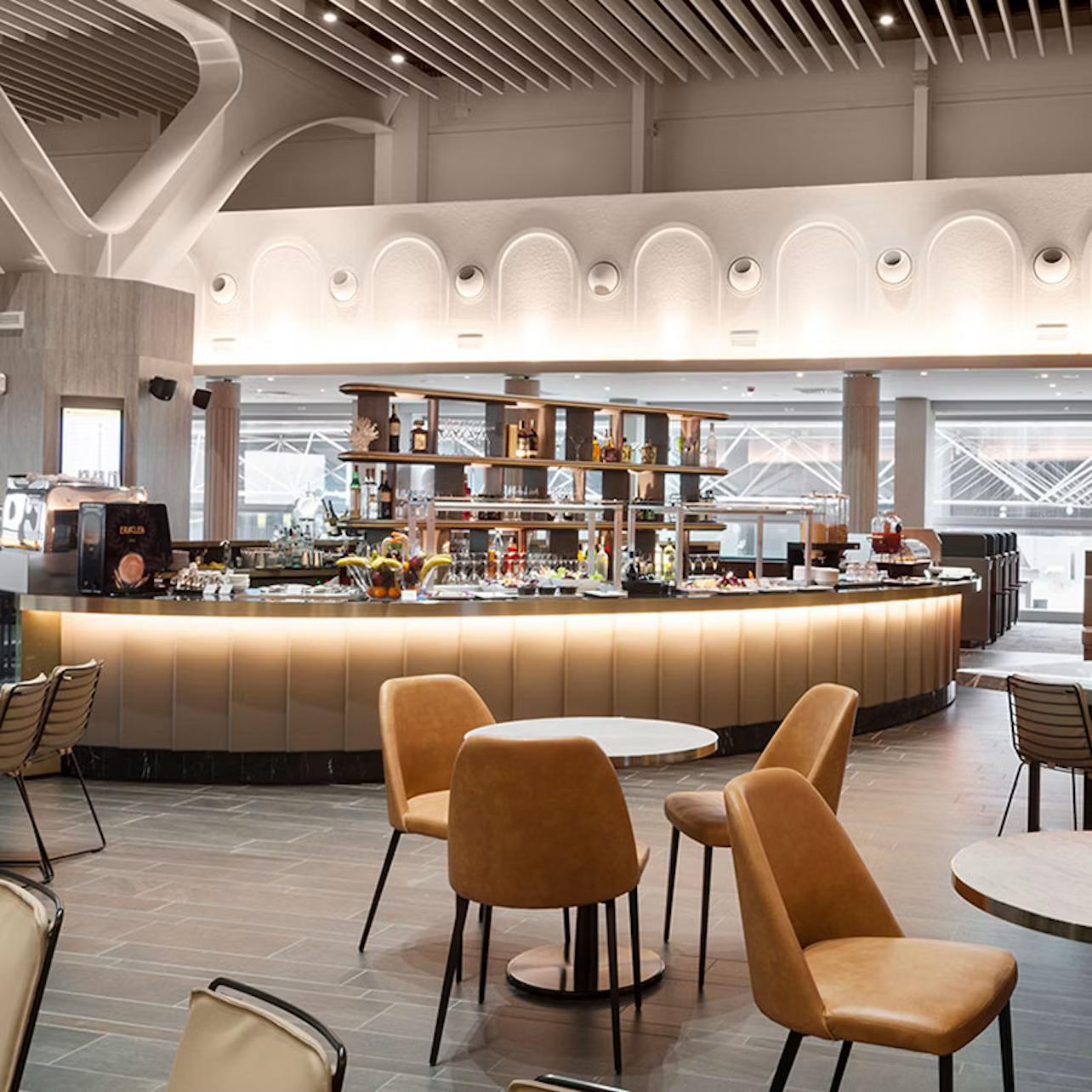 Modern bar area with illuminated counter, shelves stocked with bottles, and seating with tan chairs and round tables.