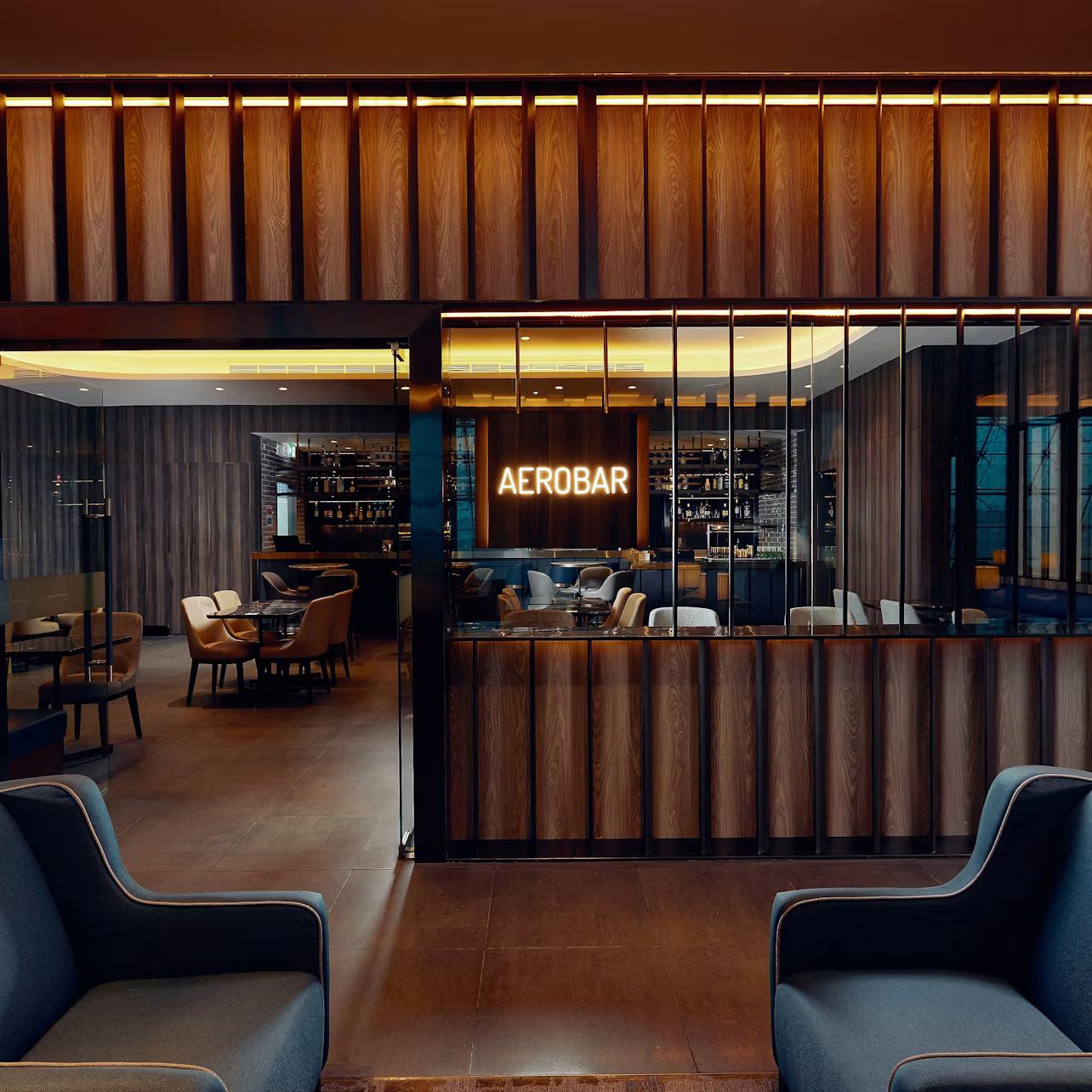 A modern bar interior with a neon sign reading "AEROBAR," wooden panels, lounge chairs, and a visible seating area with tables.